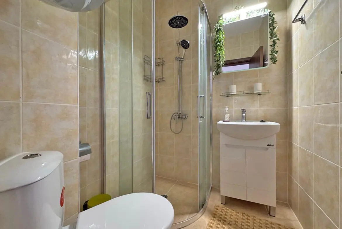 Современная ванная комната с душевой кабиной, умывальником и зелеными растениями, оформлена в светлых тонах.