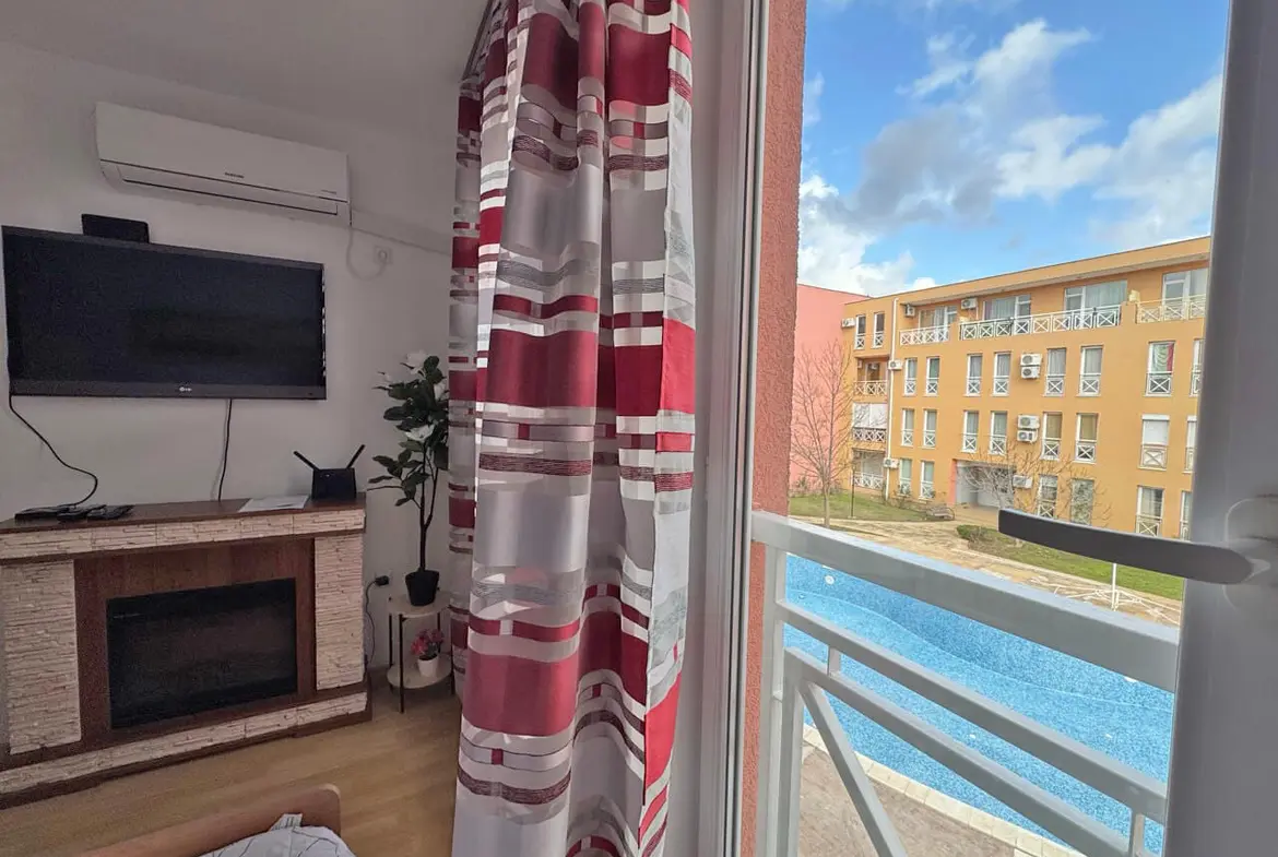 Современная студия с телевизором, диваном и видом на бассейн и жилой комплекс в Sunny Day 6. Окна с занавесками.