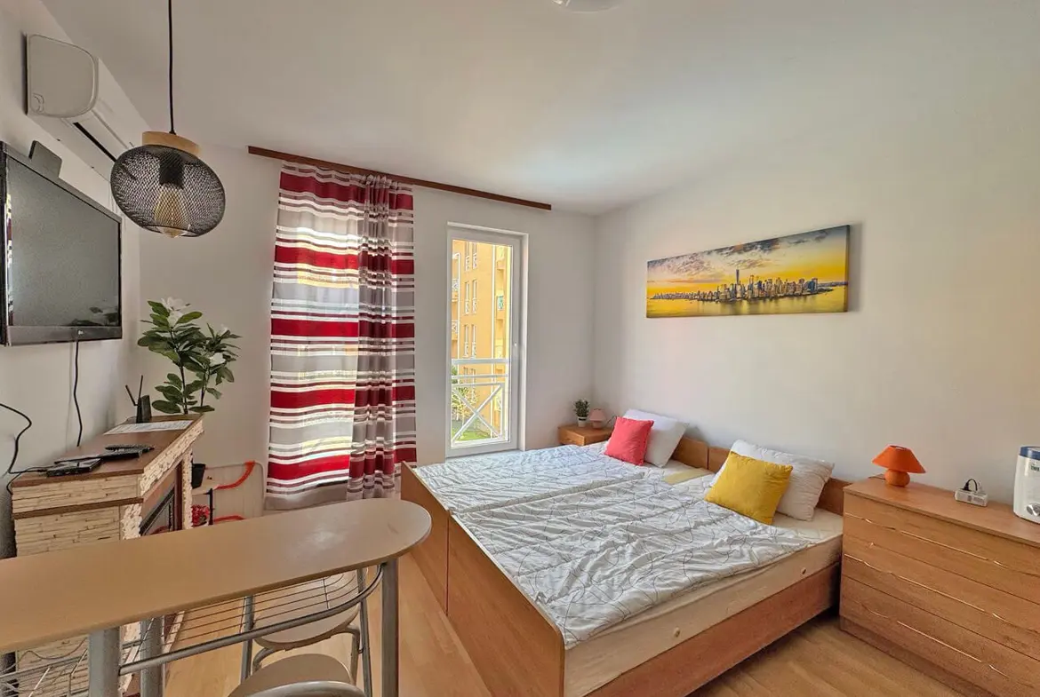 Современная студия с двуспальной кроватью, яркими подушками и телевизором, окна выходят на балкон с видом. Светлые тона интерьера.