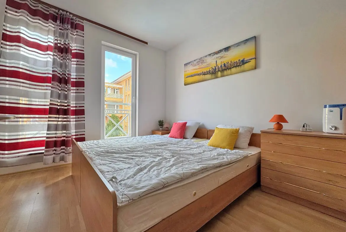 Современная спальня с двуспальной кроватью, яркими подушками, деревянной мебелью и большими окнами с рулонными шторами.