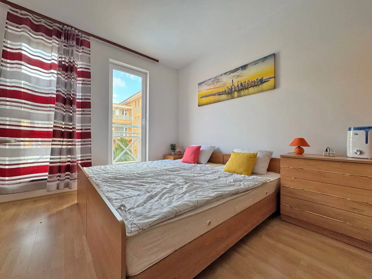 Современная спальня с двуспальной кроватью, яркими подушками, деревянной мебелью и большими окнами с рулонными шторами.