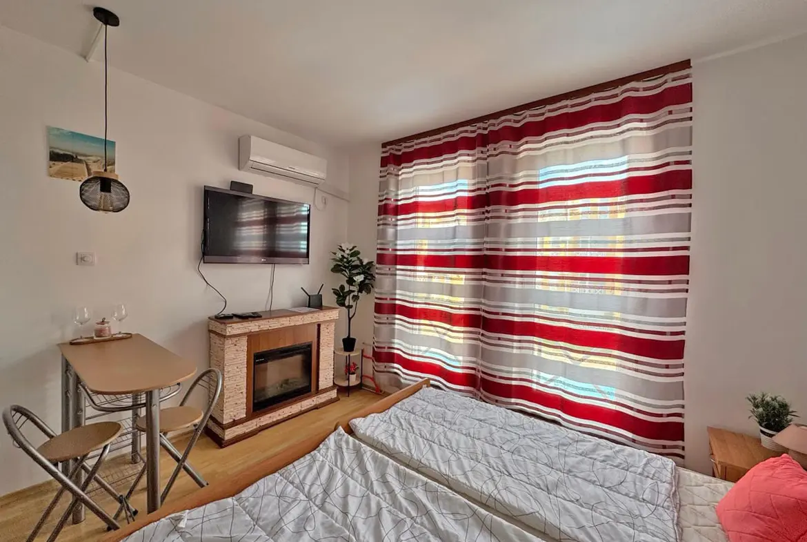 Современная студия с двуспальной кроватью, телевизором, стильным камином и уютной обеденной зоной у окна.