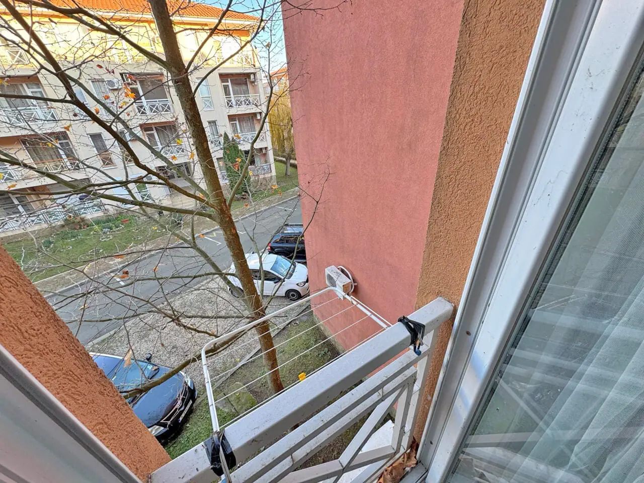 Балконная веранда с видом на улицу и припаркованными автомобилями, окружающими деревья и дома.