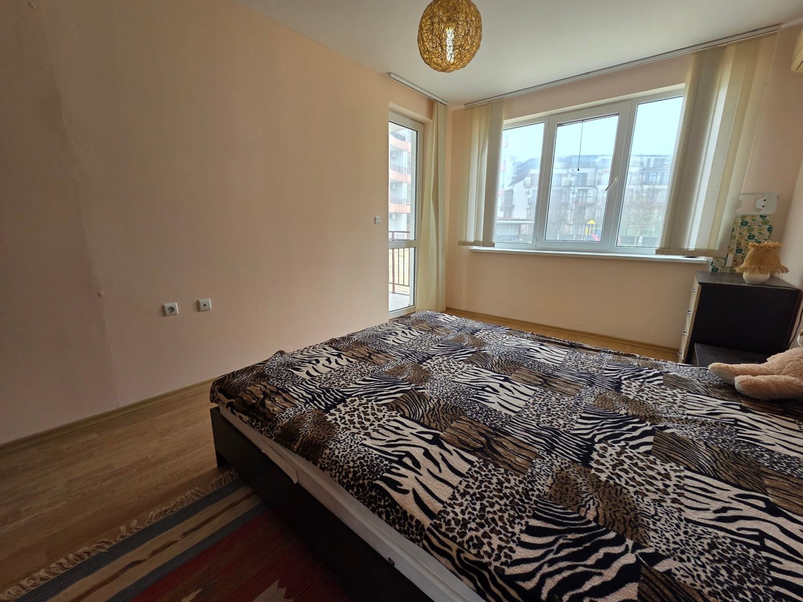 Двухкомнатный апартамент в Riviera Fort Beach – уютная спальня Современная спальня с двуспальной кроватью, светлыми стенами, панорамными окнами и стильным освещением.