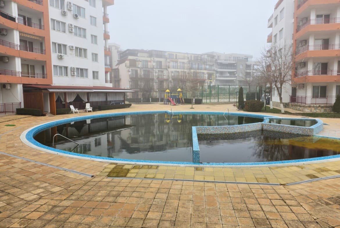 Двухкомнатный апартамент с видом на бассейн, окруженный зданиями комплекса Riviera Fort Beach, в туманной обстановке.