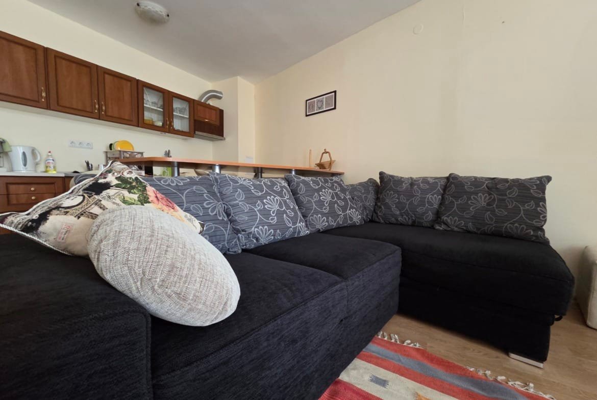 Уютный черный диван с декоративными подушками в светлом двухкомнатном апартаменте с кухней и деревянными шкафами.