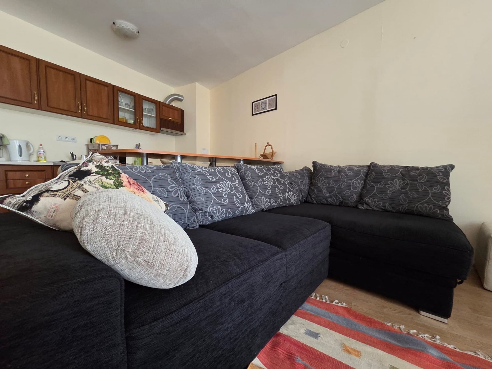 Двухкомнатный апартамент: уютный диван в Riviera Fort Beach Уютный черный диван с декоративными подушками в светлом двухкомнатном апартаменте с кухней и деревянными шкафами.