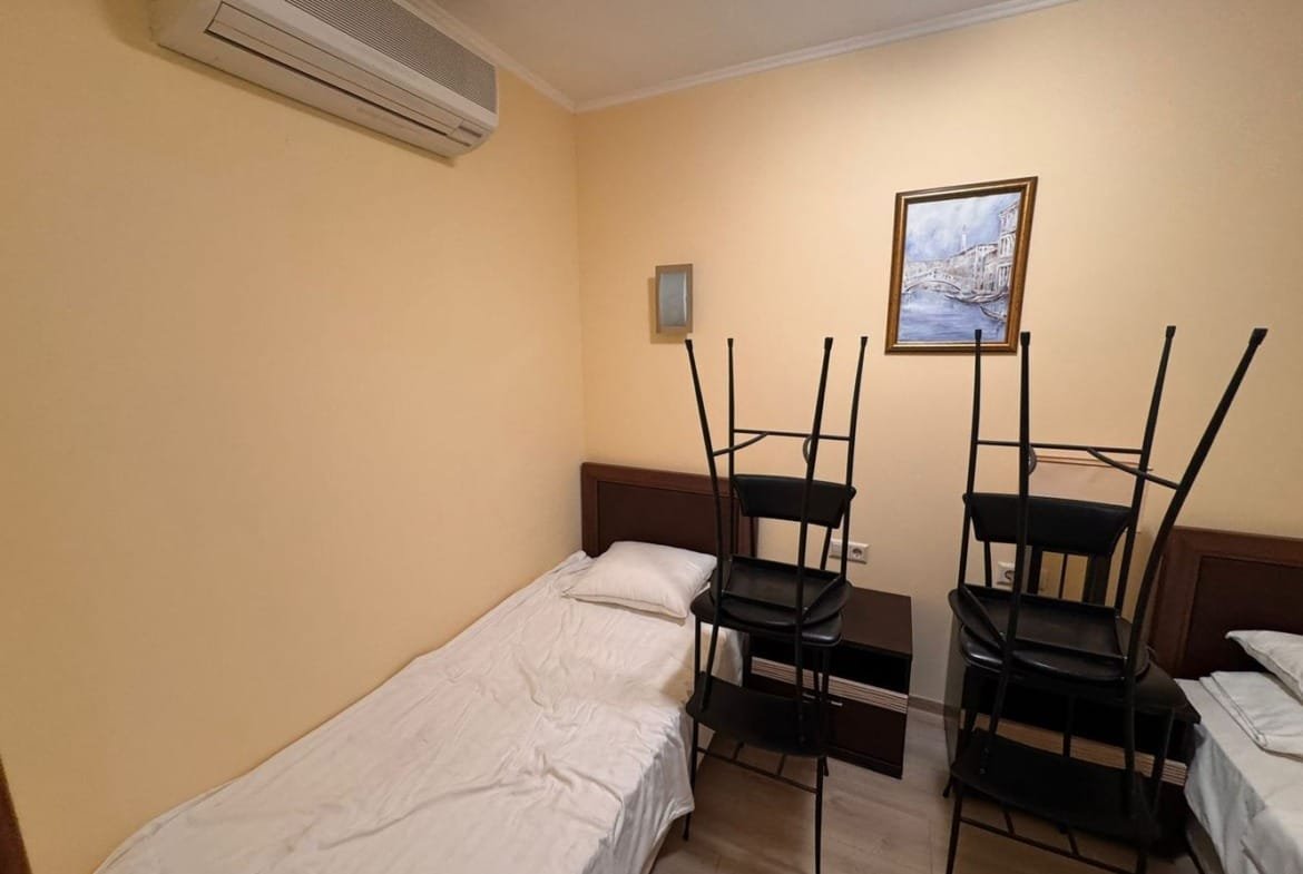 Трехкомнатный апартамент с одним спальным местом и стульями, сложенными на кровати, жёлтыми стенами и кондиционером.