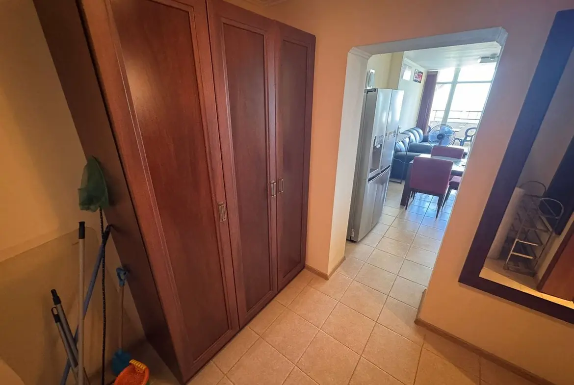 Трехкомнатный апартамент: коридор с шкафом и кухней в “Sunset Resort” Вид на коридор трехкомнатного апартамента с деревянным шкафом, ведет в гостиную с диваном и кухней.