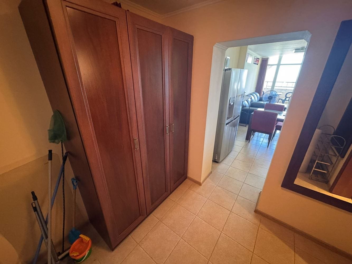 Трехкомнатный апартамент: коридор с шкафом и кухней в “Sunset Resort” Вид на коридор трехкомнатного апартамента с деревянным шкафом, ведет в гостиную с диваном и кухней.