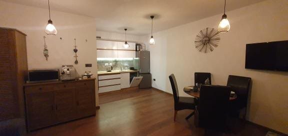 Современная студия с кухней, обеденным столом и телевизором, оформленная в теплых тонах, на деревянном полу.