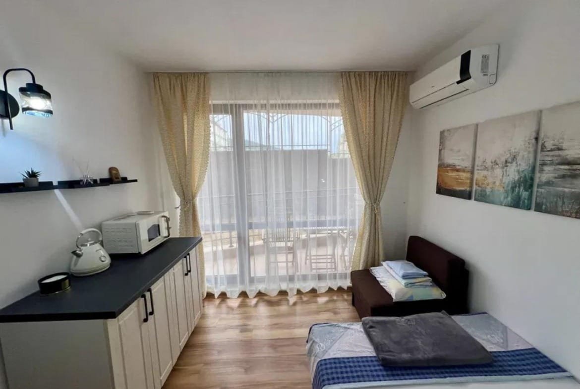 Уютная студия с современной мебелью, окнами, светлыми шторами и небольшим кухонным уголком в комплексе “Macon Residence”.