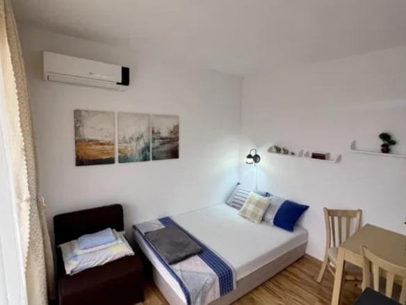 Современная студия с односпальной кроватью, столом и стульями, оформленная в светлых тонах, с деревянным полом.