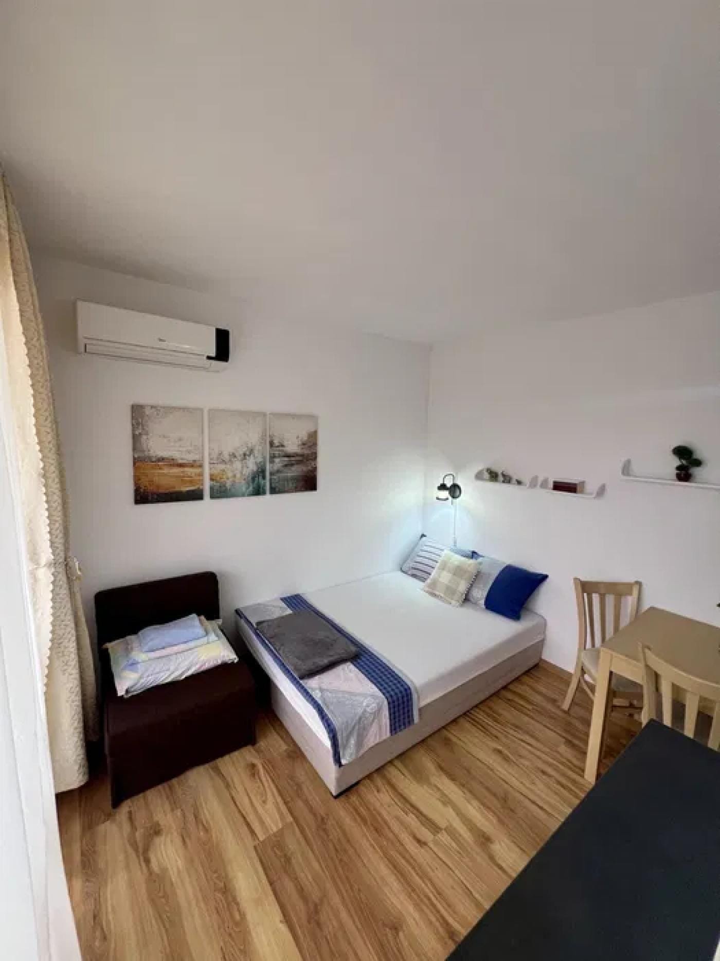 Современная студия с односпальной кроватью, столом и стульями, оформленная в светлых тонах, с деревянным полом.