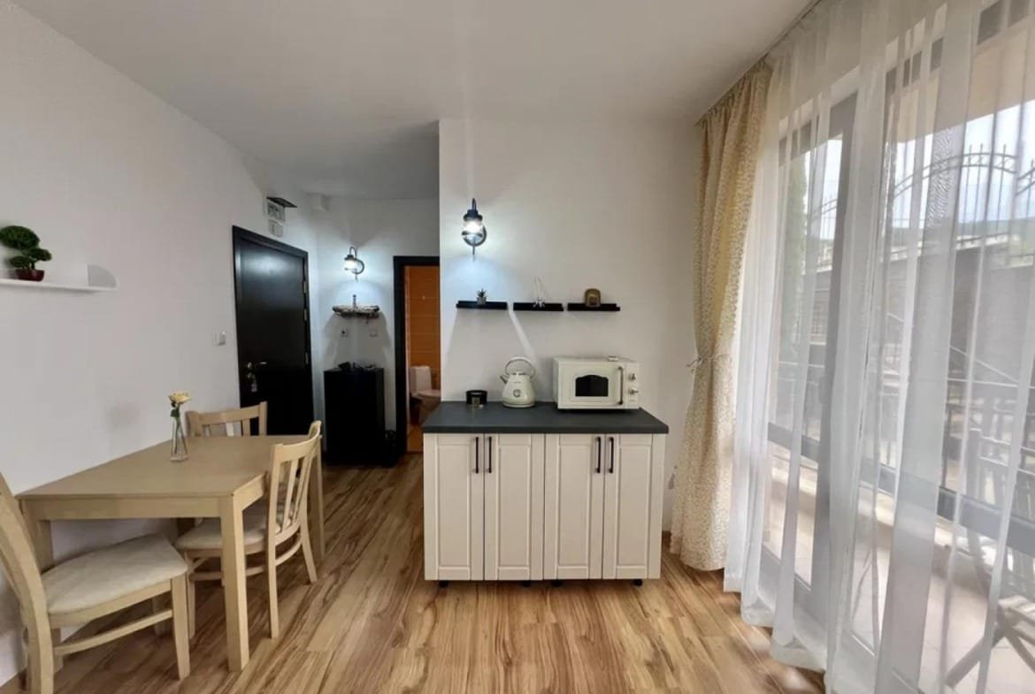 Современная студия с кухонным уголком, столом, стульями и балконной дверью, оформленная в светлых тонах.