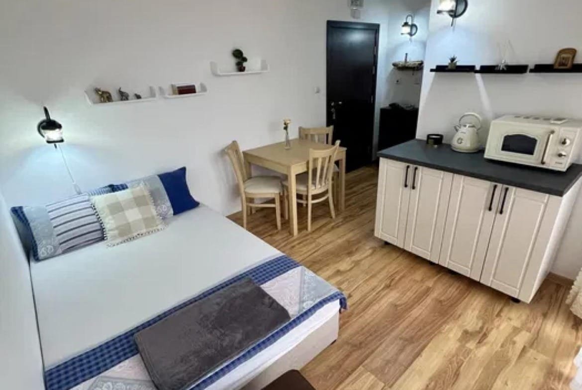 Современная студия с двуспальной кроватью, кухней и обеденным столом, оформленная в светлых тонах.