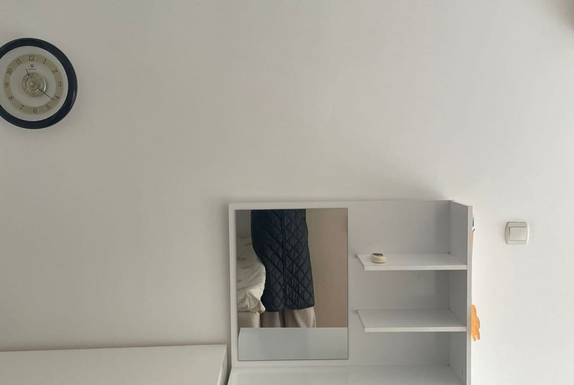 Современный комод с зеркалом и полками на белой стене, рядом с часами и креслом, оформление в стиле минимализма.