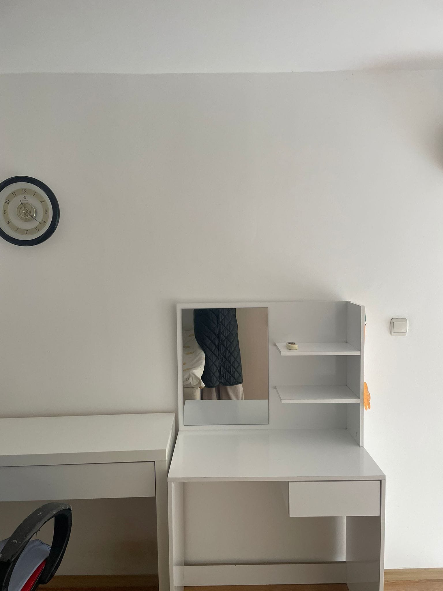 Современный комод с зеркалом и полками на белой стене, рядом с часами и креслом, оформление в стиле минимализма.