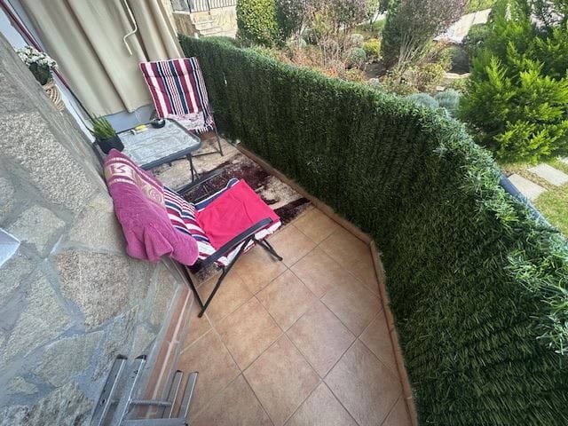 Балкон с зелёным забором, креслами и столом, украшенный ковром и полотенцами, создает уютную атмосферу для отдыха.