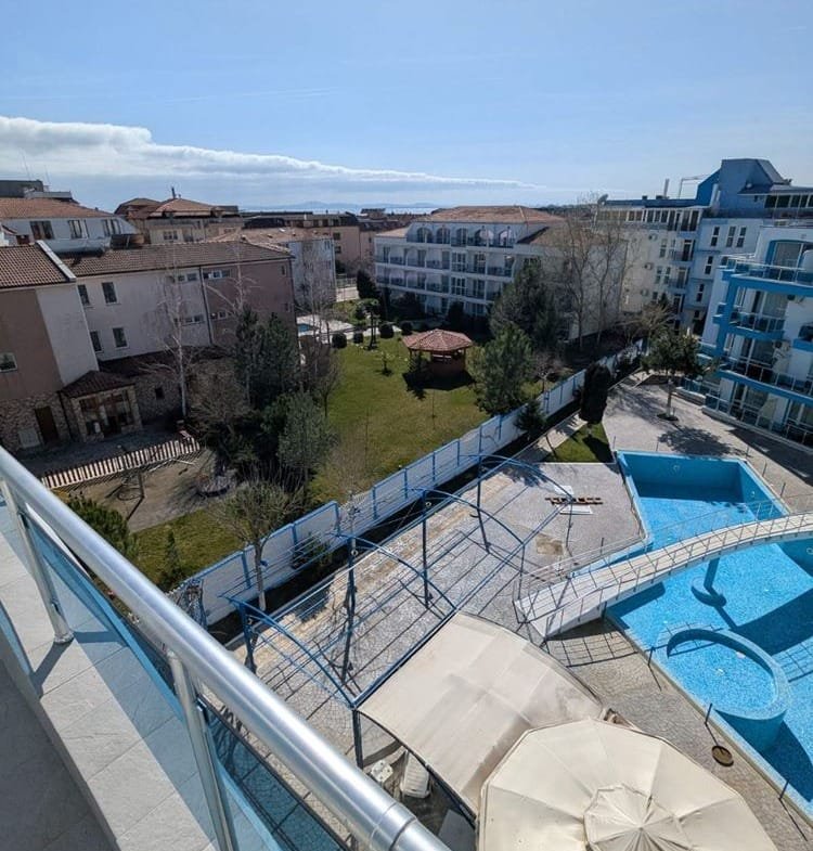 Вид с балкона на зелёный двор комплекса Odyssey, крытый бассейн и жилые здания под ясным небом.