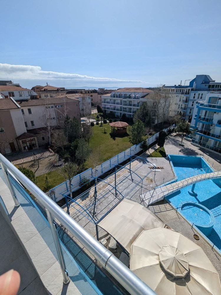 Вид с балкона на зелёный двор комплекса Odyssey, крытый бассейн и жилые здания под ясным небом.