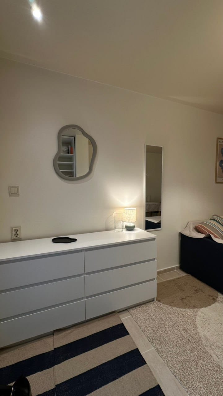 Современный интерьер студии с белым комодом, зеркалом и уютным текстилем на диване. Светлые тона и минимализм.