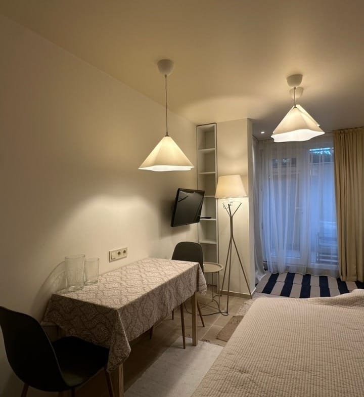 Современная студия с обеденным столом, телевизором и светлыми занавесками, уютная атмосфера, нейтральные тона.