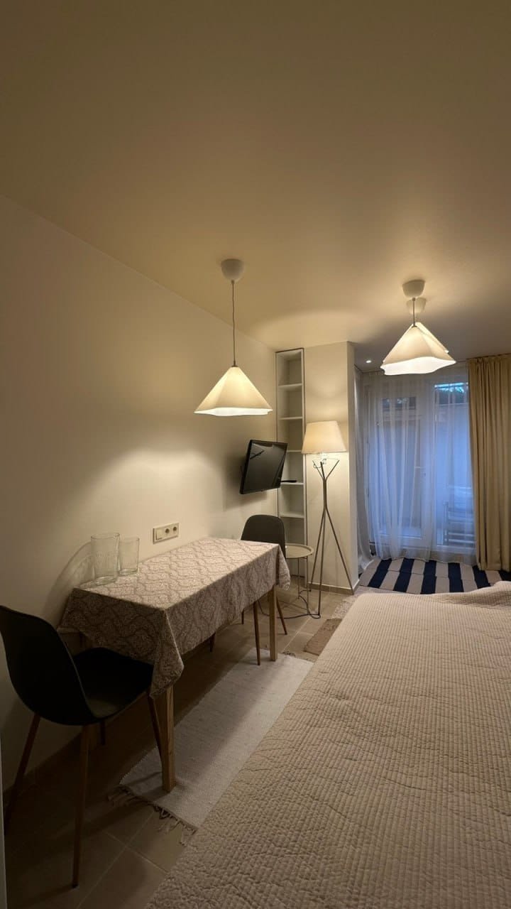 Современная студия с обеденным столом, телевизором и светлыми занавесками, уютная атмосфера, нейтральные тона.