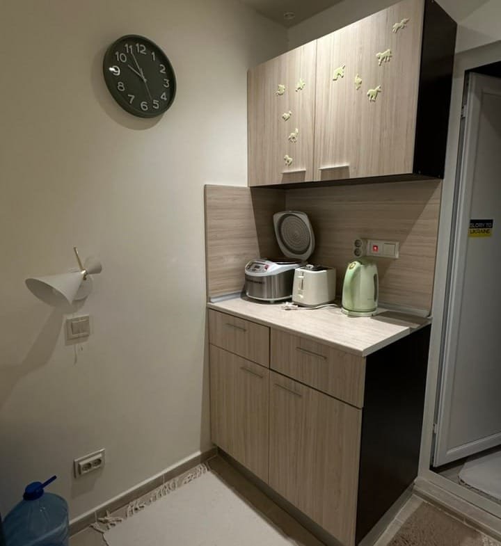 Современная кухня в студии с деревянными шкафами, плитой, чайником и часами на стене, освещённая мягким светом.