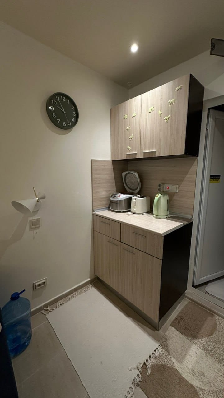 Современная кухня в студии с деревянными шкафами, плитой, чайником и часами на стене, освещённая мягким светом.