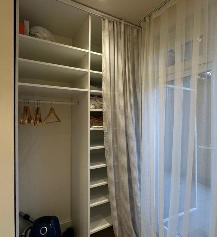 Современный гардероб с полками и вешалками, рядом пылесос, легкие занавески и нейтральные тона.