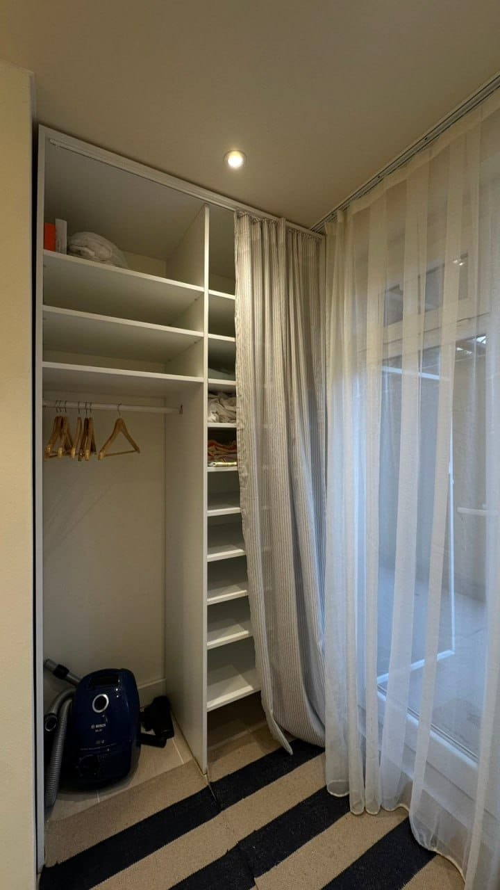 Современный гардероб с полками и вешалками, рядом пылесос, легкие занавески и нейтральные тона.