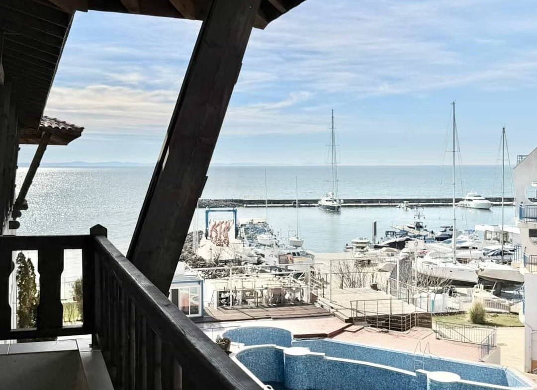 Вид с балкона на марину с яхтами и спокойное море, светлое небо и солнечные отражения на воде.