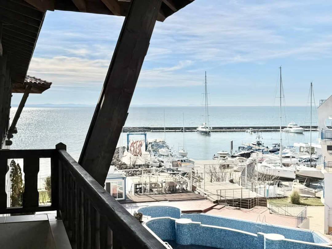 Вид с балкона на марину с яхтами и спокойное море, светлое небо и солнечные отражения на воде.