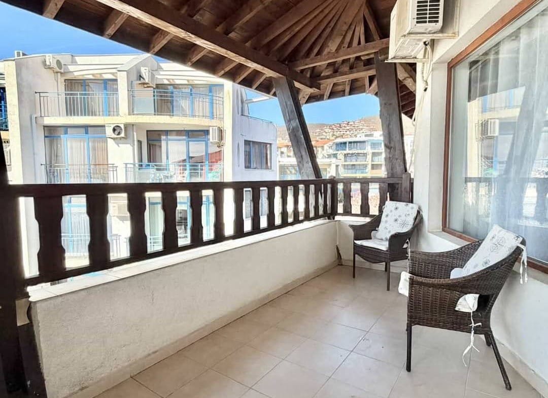 Уютный балкон с креслами и видом на здания комплекса, открытая терраса с деревянной крышей и светлыми плитками.