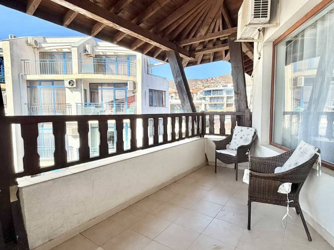 Уютный балкон с креслами и видом на здания комплекса, открытая терраса с деревянной крышей и светлыми плитками.