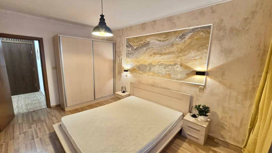 Современная спальня с двуспальной кроватью, светлым шкафом и картиной на стене в теплых тонах. Домашняя атмосфера уюта.