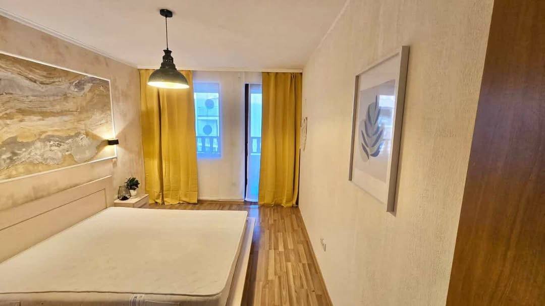 Современная спальня с светлыми стенами, лампой над кроватью, желтыми шторами и деревянным полом.