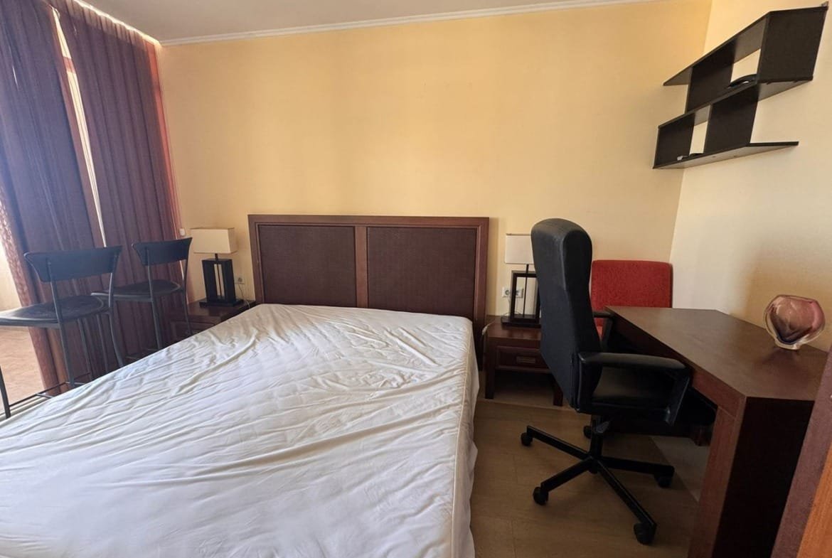 Светлая комната с кроватью без постельного белья, столом, стулом и барными стульями у окна. Просторный интерьер с нейтральными оттенками.