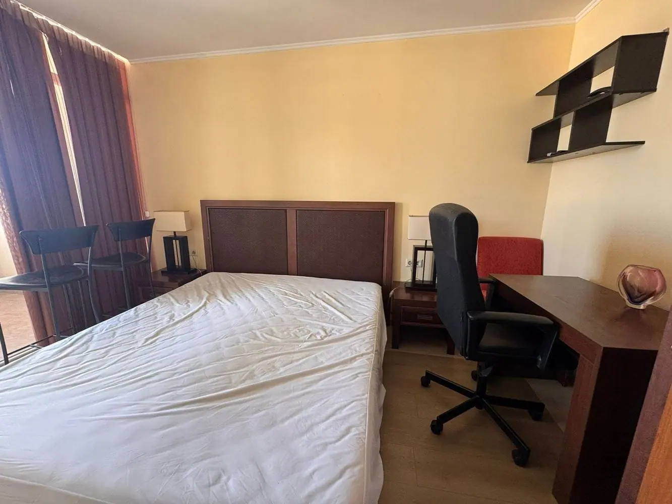 Современный интерьер трехкомнатного апартамента в “Sunset Resort” Светлая комната с кроватью без постельного белья, столом, стулом и барными стульями у окна. Просторный интерьер с нейтральными оттенками.