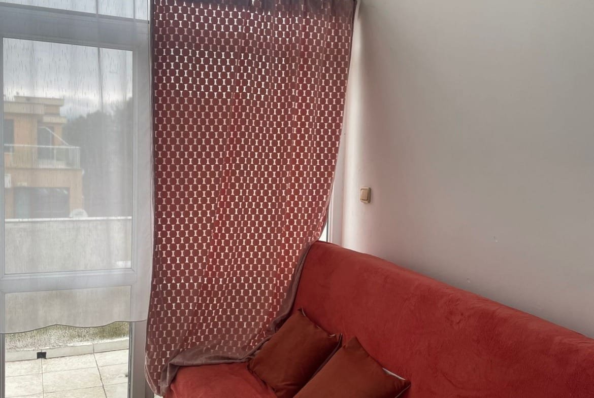 Уютная комната с красным диваном и занавеской, света много и есть выход на балкон. Настенные часы на фоне.