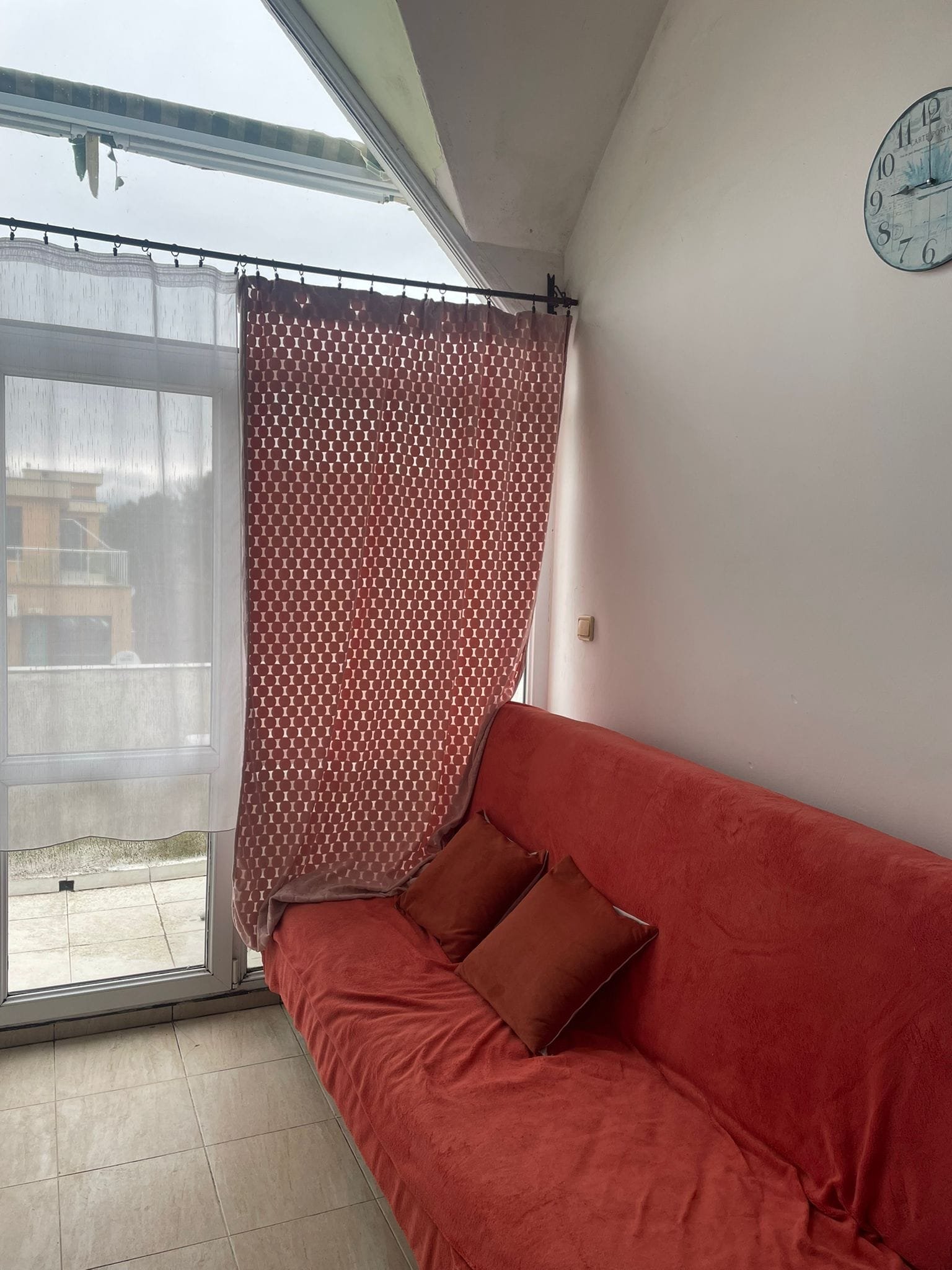 Уютная комната с красным диваном и занавеской, света много и есть выход на балкон. Настенные часы на фоне.