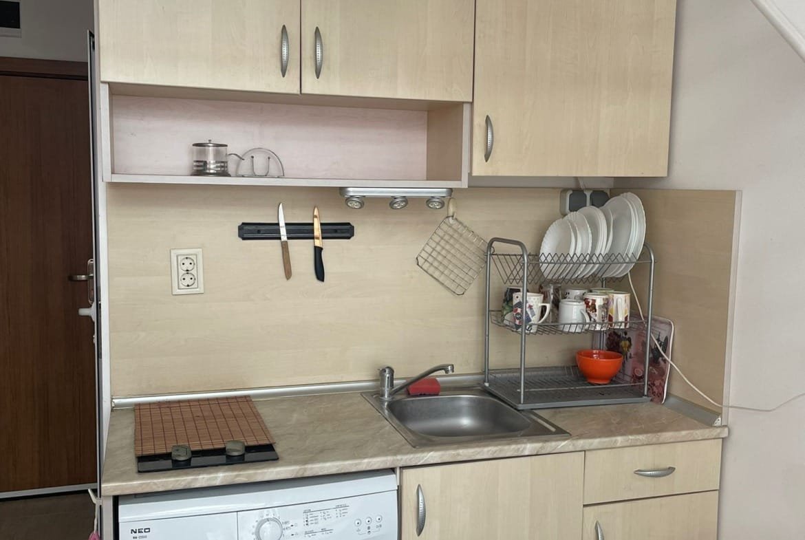 Современная кухня в студии с мойкой, плитой, стиральной машиной и открытыми полками для посуды. Светлые тона, уютная атмосфера.