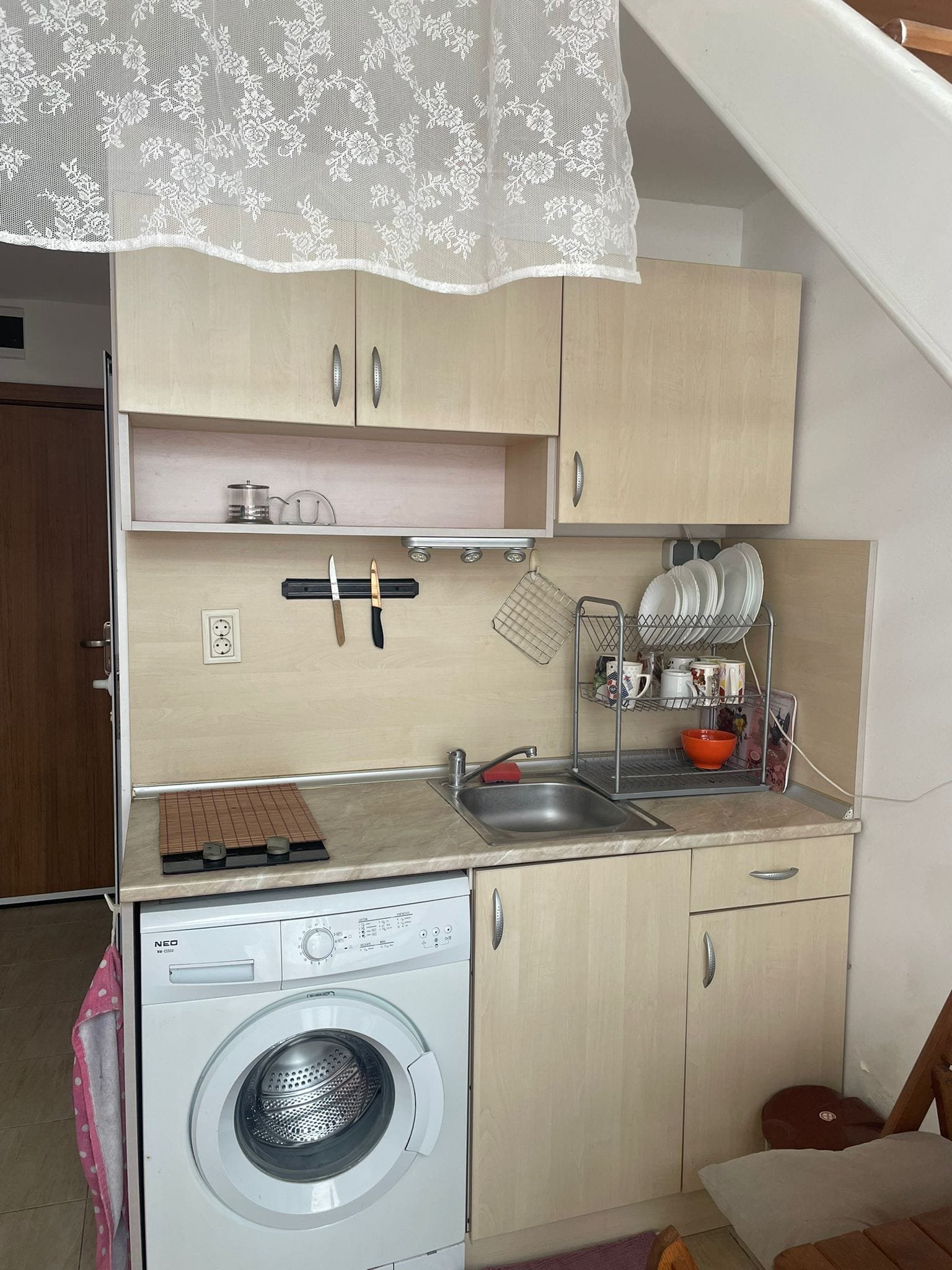 Современная кухня в студии с мойкой, плитой, стиральной машиной и открытыми полками для посуды. Светлые тона, уютная атмосфера.