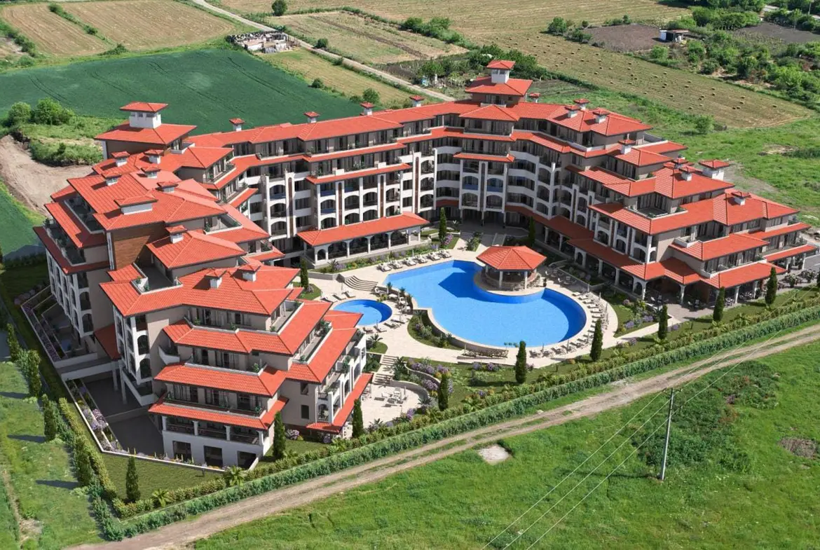 Жилой комплекс с красной крышей, бассейном и landscaped территорией, окружённый зелёными полями.