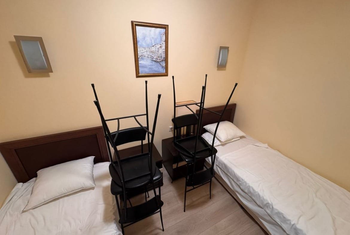 Две кровати с пустыми постелями, между ними стол, на котором стоят складывающиеся стулья, стены светло-желтого цвета.