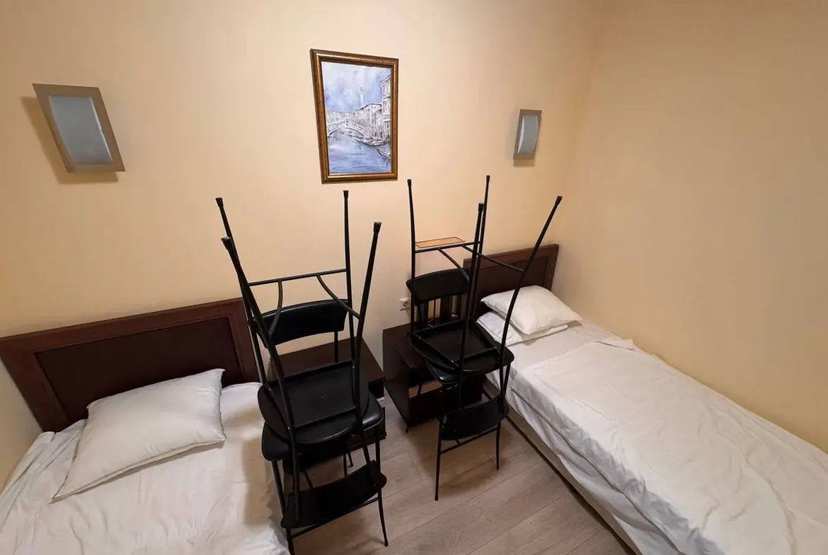 Трехкомнатный апартамент в “Sunset Resort” с двумя кроватями. Две кровати с пустыми постелями, между ними стол, на котором стоят складывающиеся стулья, стены светло-желтого цвета.