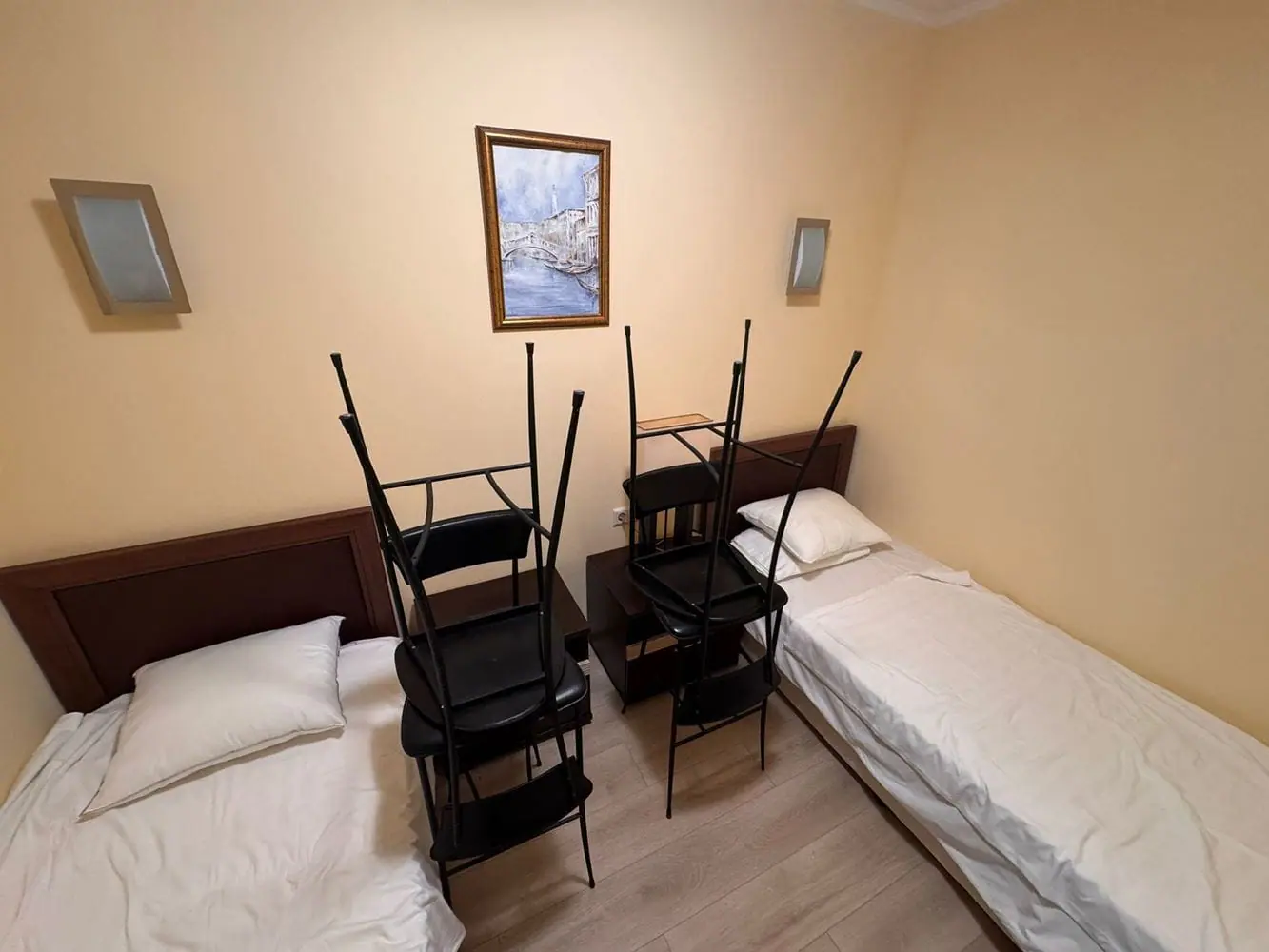 Трехкомнатный апартамент в “Sunset Resort” с двумя кроватями. Две кровати с пустыми постелями, между ними стол, на котором стоят складывающиеся стулья, стены светло-желтого цвета.