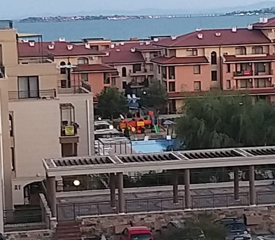 Вид на жилой комплекс с детской площадкой и бассейном, расположенный рядом с морем и зелеными насаждениями.