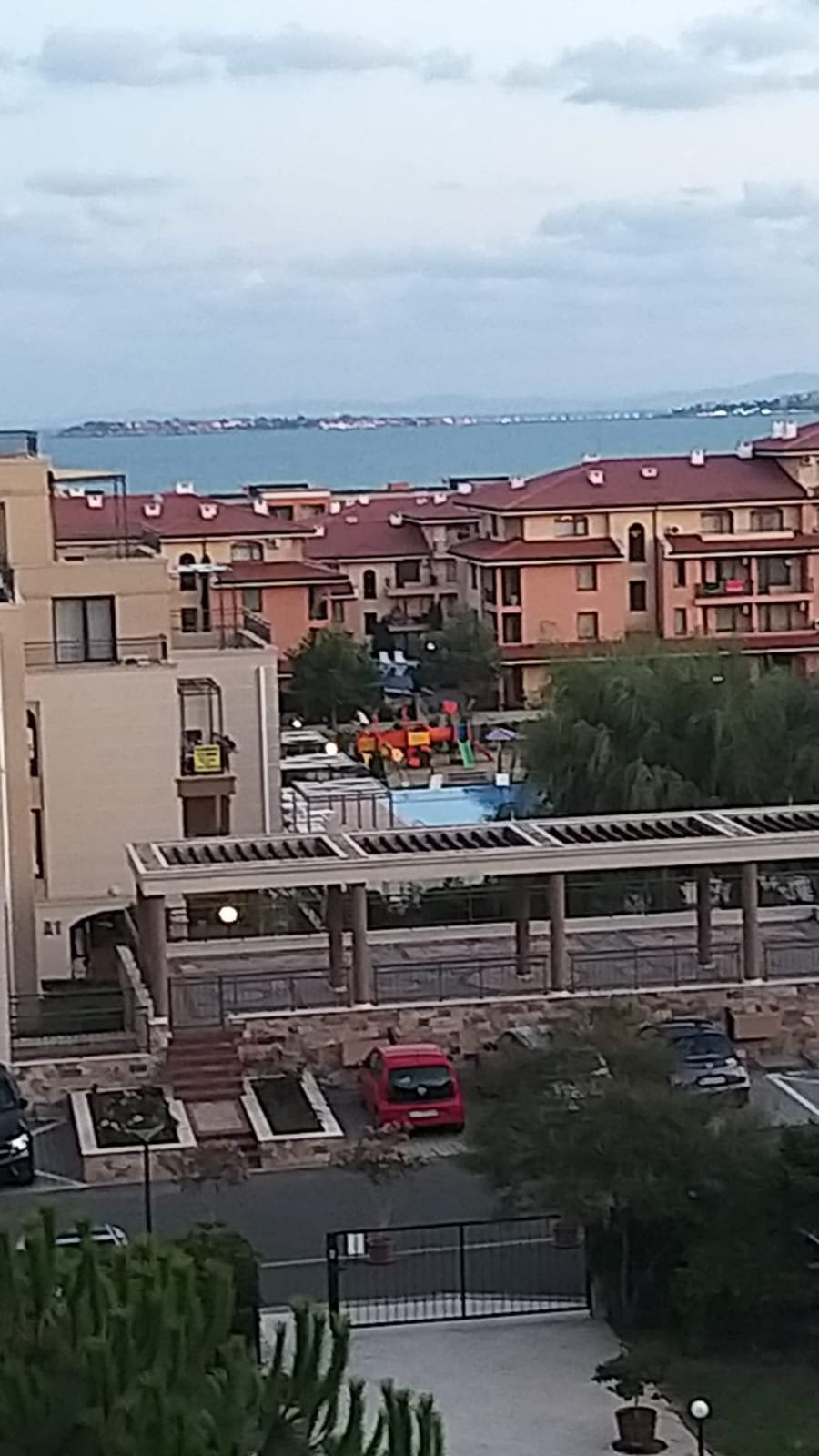 Вид на жилой комплекс с детской площадкой и бассейном, расположенный рядом с морем и зелеными насаждениями.